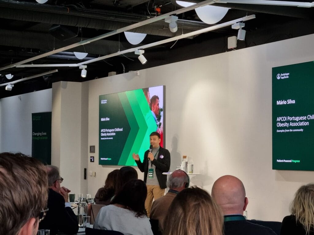 A speaker presents at a conference, standing in front of a screen displaying text and graphics, with an audience seated in front.
