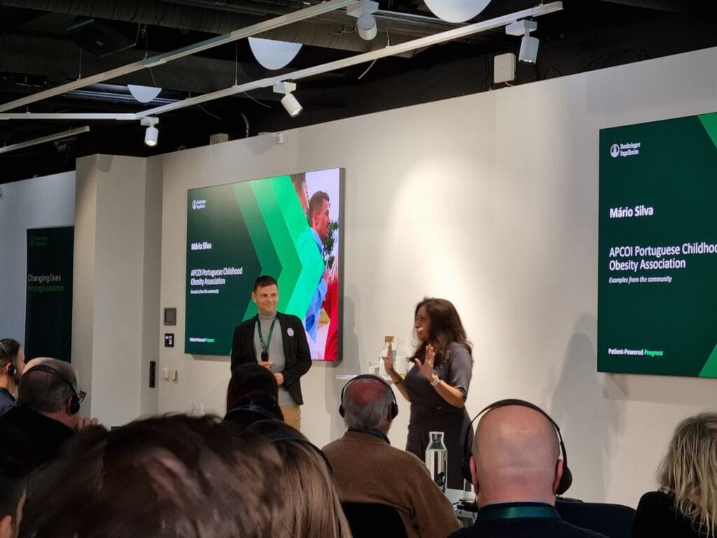 Two speakers stand in front of an audience in a conference room. One man in a suit smiles while a woman gestures as she speaks. Behind them are presentation screens and banners.