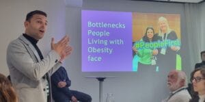 A person giving a presentation in a conference room. The slide reads "Bottlenecks People Living with Obesity face.