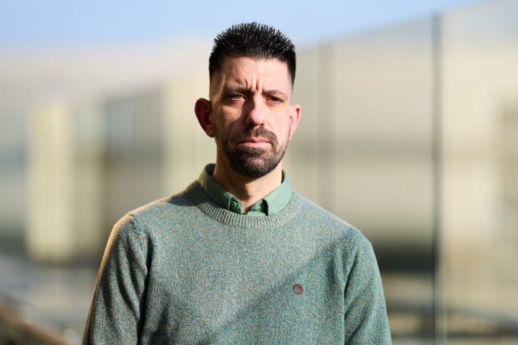 A man with short dark hair and a beard stands outdoors, wearing a green sweater over a collared shirt. Blurred buildings are visible in the background.