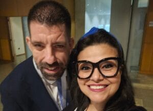 Two people are posing for a selfie in an indoor setting. The woman is smiling and wearing glasses with a white shirt, while the man is dressed in a suit.