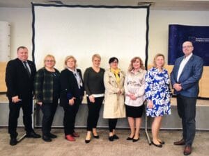 Eight adults stand in a row in front of a blank projection screen indoors, dressed in business or semi-formal attire.