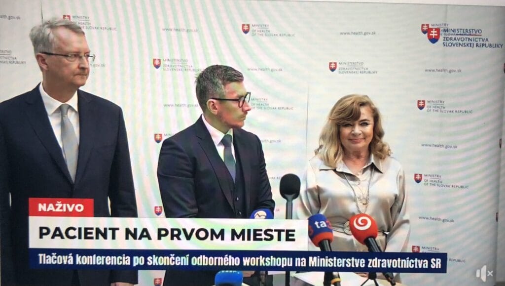 Three people stand at a press conference podium in front of a Ministry of Health backdrop, with microphones and news logos visible in the foreground.