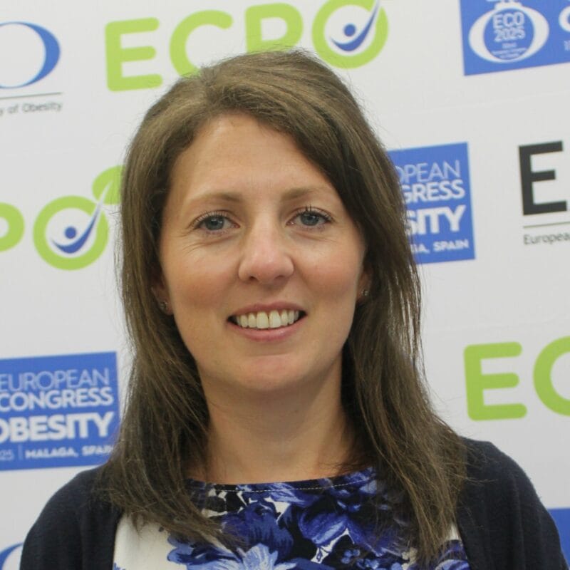A woman with brown hair smiles at the camera, standing in front of a backdrop with logos for the European Congress on Obesity and ECPO.