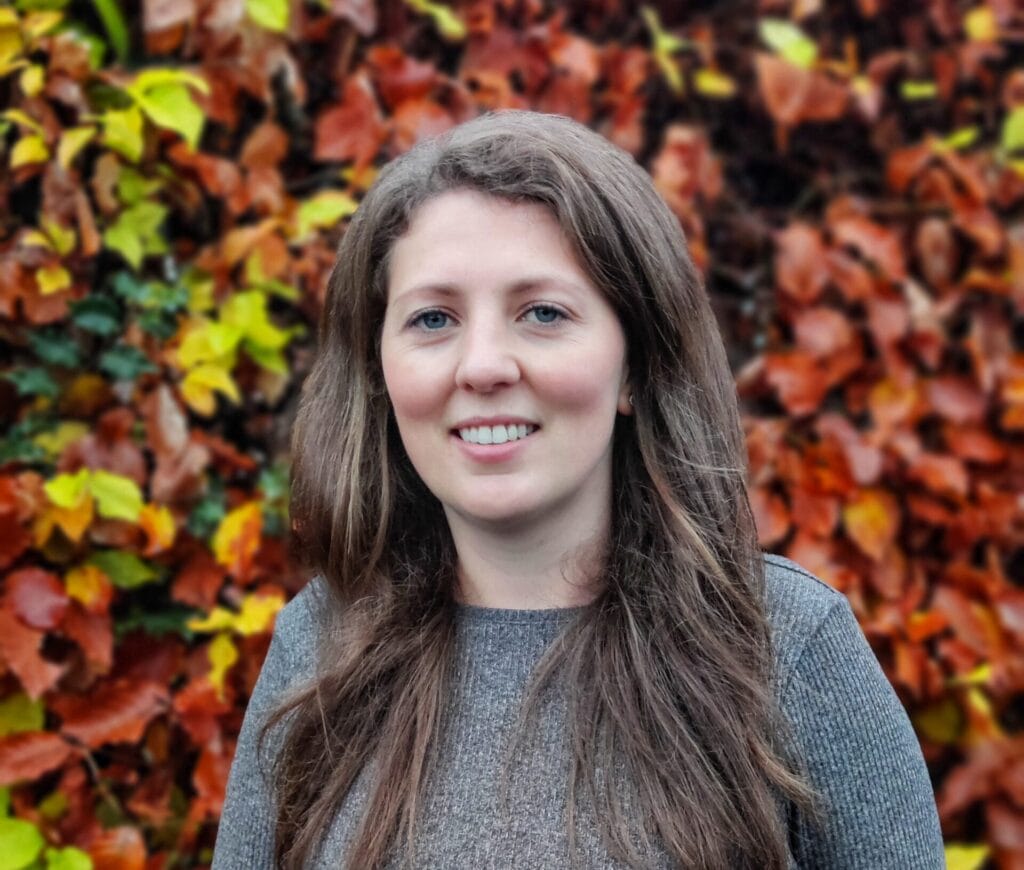 Person with long brown hair wearing a gray sweater standing in front of a vibrant background of autumn leaves.