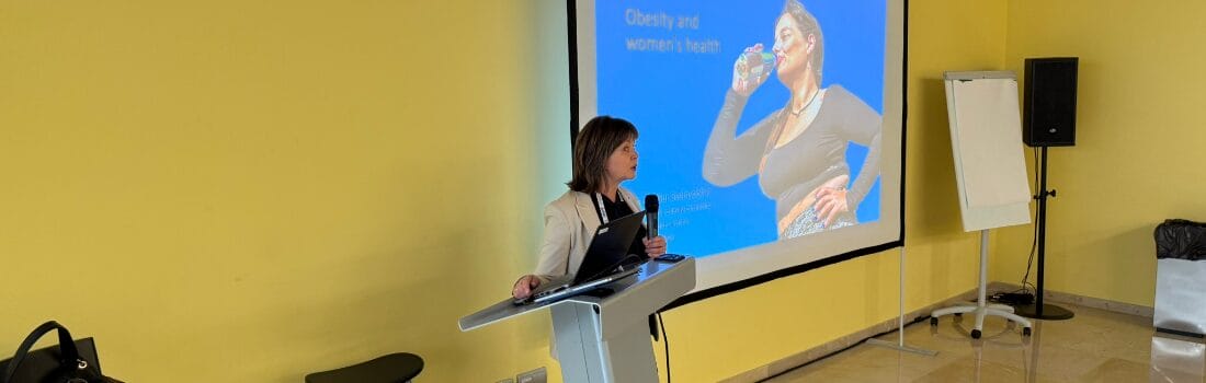 A woman stands at a podium giving a presentation on "Obesity and women’s health," with a projection screen showing a woman drinking water.