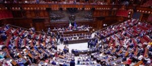 Large parliamentary chamber with many people seated at desks, officials at the front, and Italian and European Union flags displayed behind the main podium.