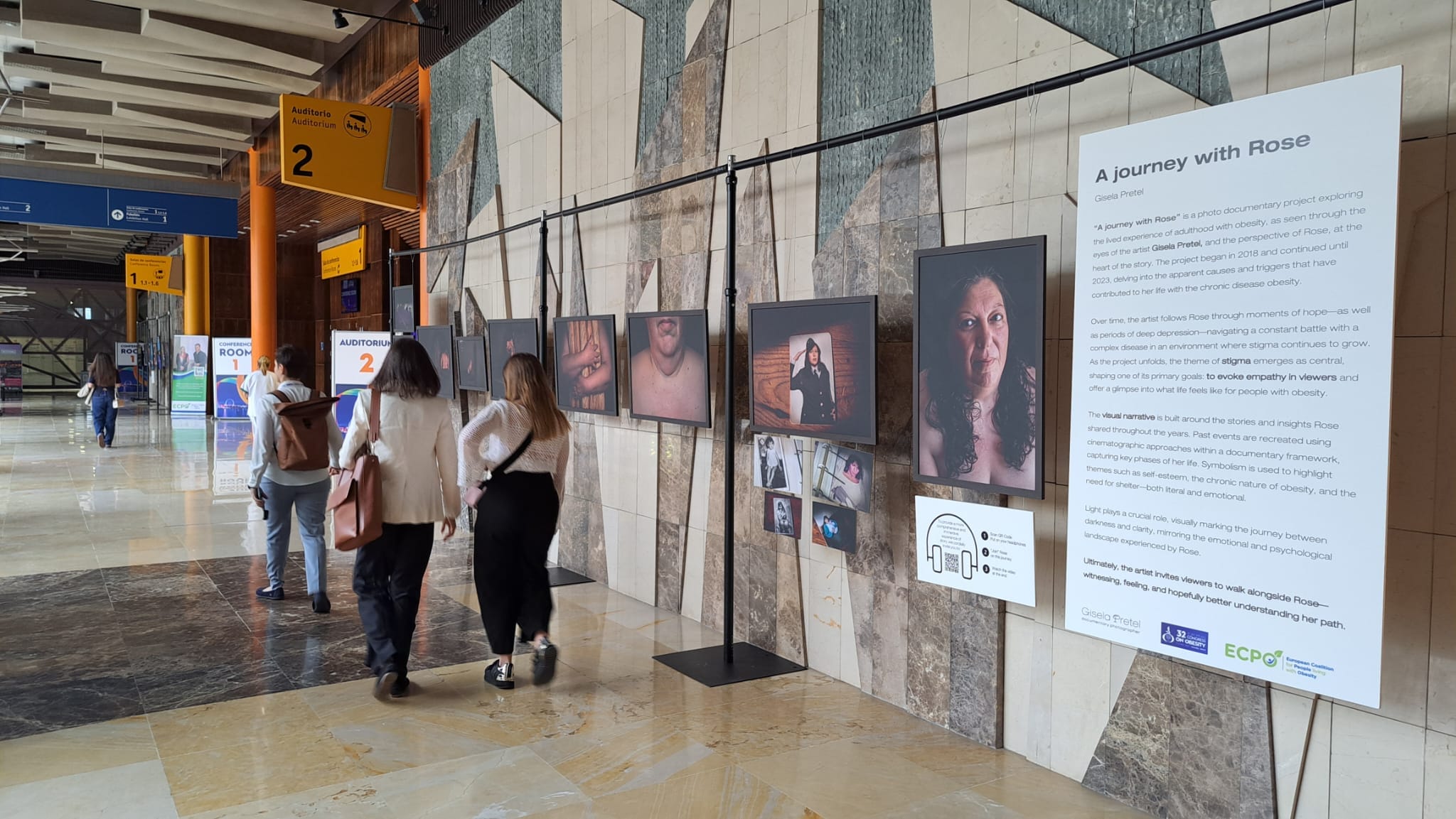 Three people walk past a photo exhibition titled “A journey with Rose” displayed on a marble wall in a public indoor space with directional signs overhead.