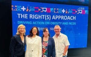 Four people stand in front of a large screen displaying the text, "THE RIGHT(S) APPROACH: DRIVING ACTION ON OBESITY AND NCDS.