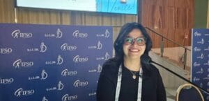 A woman stands at a podium, smiling, with a screen behind her displaying information about the 13th IFSO-EC Congress in Venice, Italy, held from 14-17 May 2025.