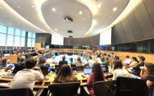A large group of people sit around a circular conference table in a modern meeting room, with laptops and papers in front of them.