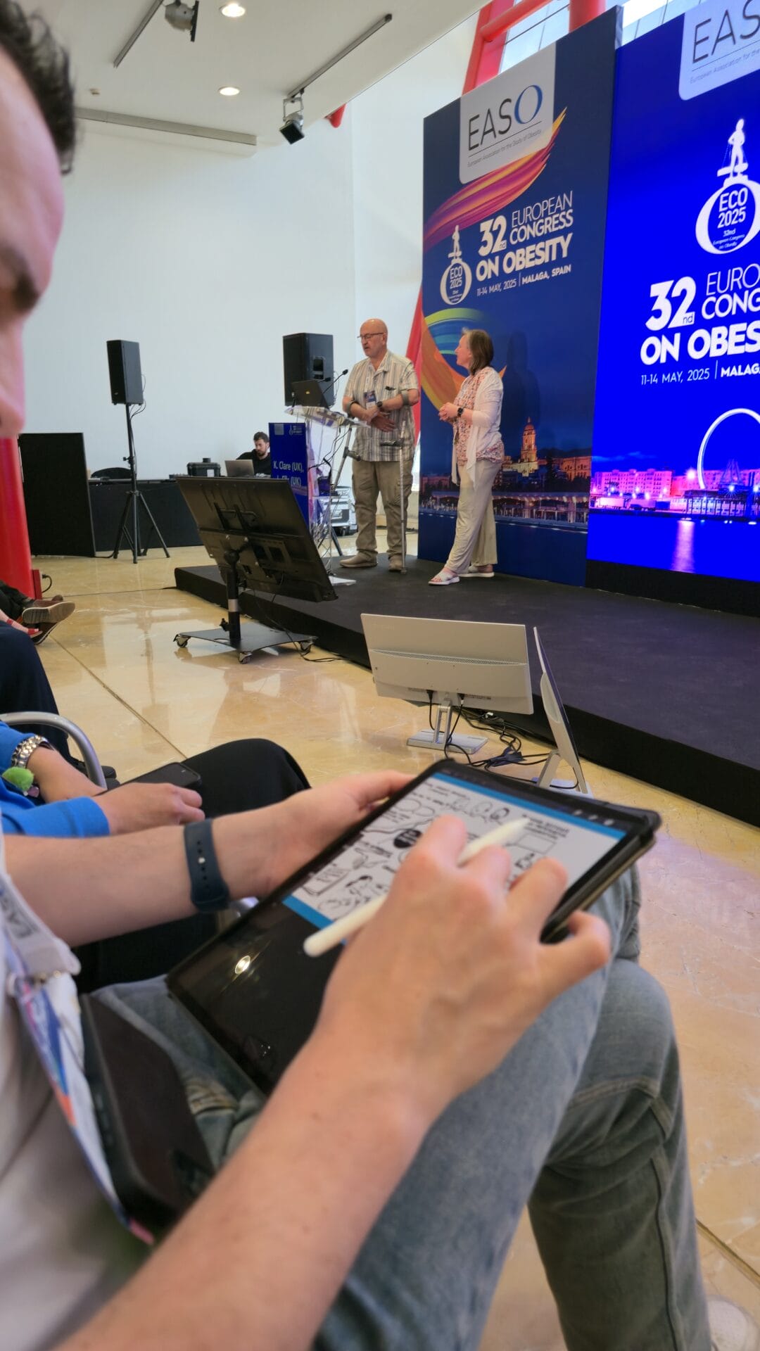 A person draws on a tablet in the audience while two speakers stand on stage at the European Congress on Obesity, with event signage in the background.