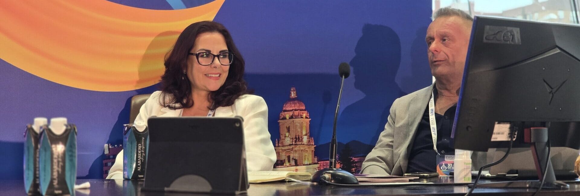 Two people sit at a conference table with electronic devices, in front of a colorful backdrop featuring an architectural image.