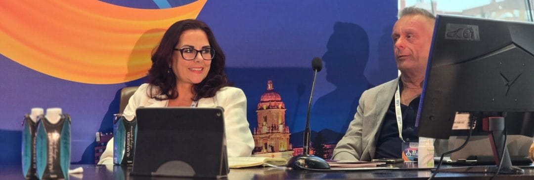 Two people sit at a conference table with electronic devices, in front of a colorful backdrop featuring an architectural image.