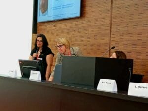 Three women are seated at a conference panel. One stands and speaks into a microphone, while another uses a computer. Name placards are visible in front of them.