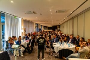 A speaker addresses a seated audience in a modern conference room, with round tables, bottled water, and presentation equipment visible.