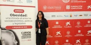 A woman stands next to a sign about an obesity conference, with university and sponsor logos in the background at an indoor event.