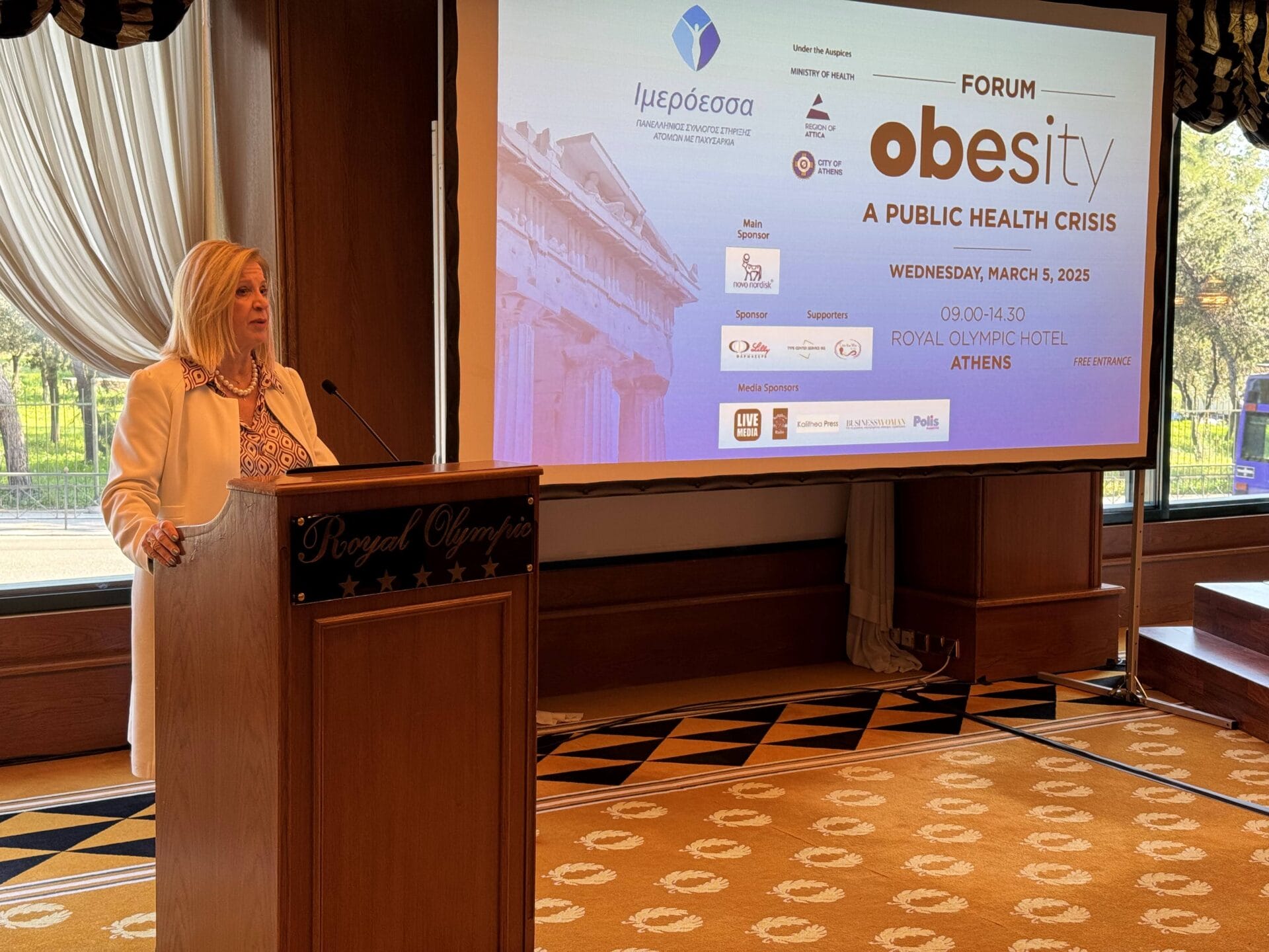 A woman stands at a podium in a conference room, speaking at the “Obesity: A Public Health Crisis” forum held at the Royal Olympic Hotel in Athens on March 5, 2025.