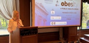 A woman speaks at a podium in a conference room, with a large screen behind her displaying information about a forum on obesity as a public health crisis in Athens.
