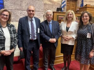 Five professionally dressed adults pose and smile for a photo inside a formal government building with wooden podiums and a Greek flag visible in the background.