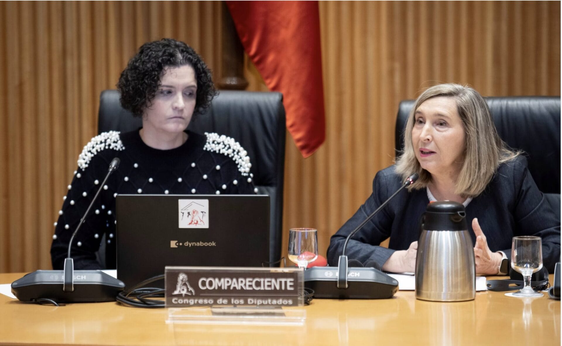 Two women sit at a conference table with microphones, a laptop, and a nameplate reading "Compareciente Congreso de los Diputados" in front of them. Two women sit at a conference table with microphones, a laptop, and a nameplate reading "Compareciente Congreso de los Diputados" in front of them.