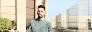 A man with short dark hair and a beard stands outdoors on a wooden deck, holding a railing, with buildings and glass panels in the background.