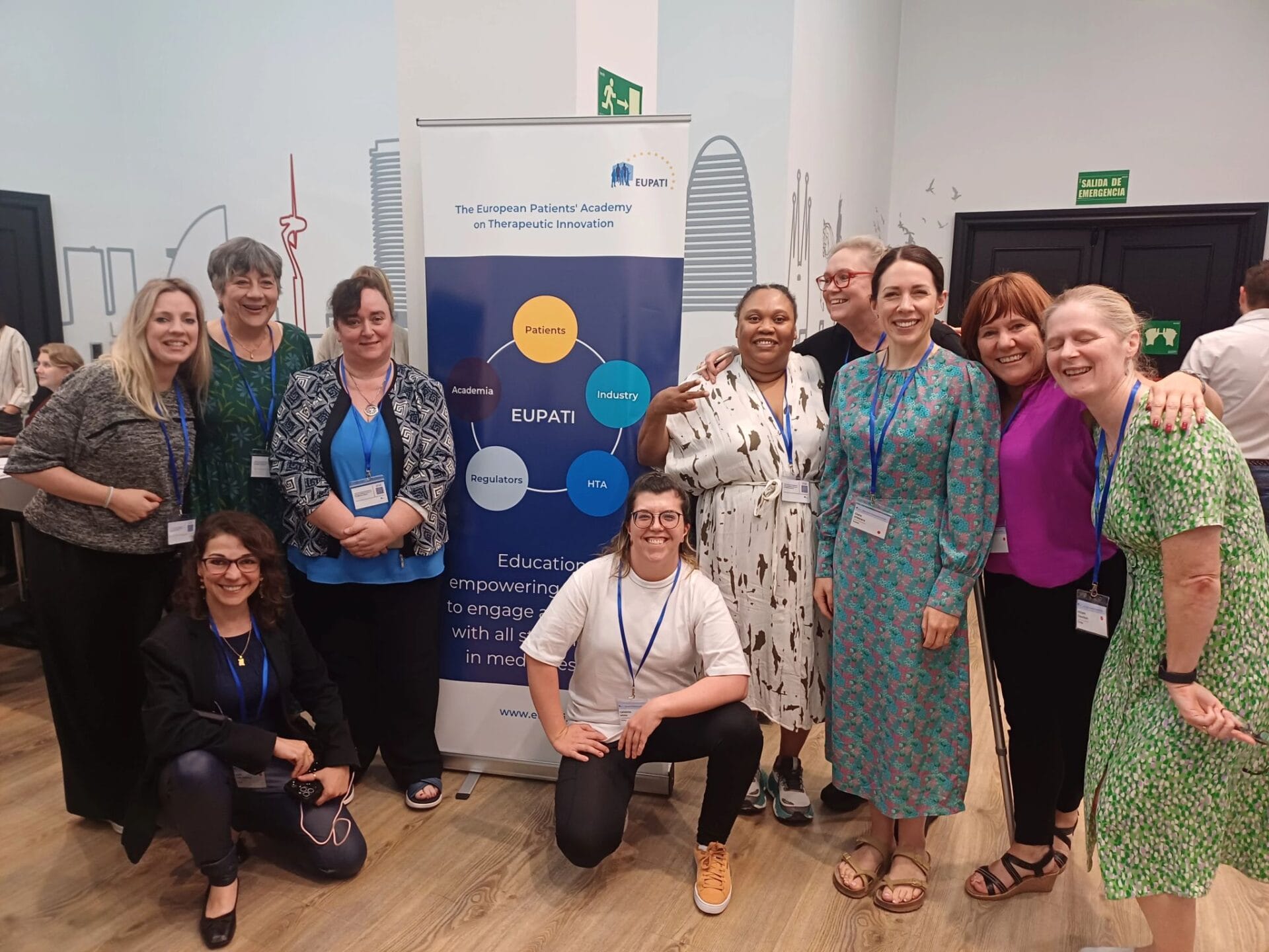 A group of people pose and smile in front of an EUPATI banner at an indoor event, with some standing and others kneeling.