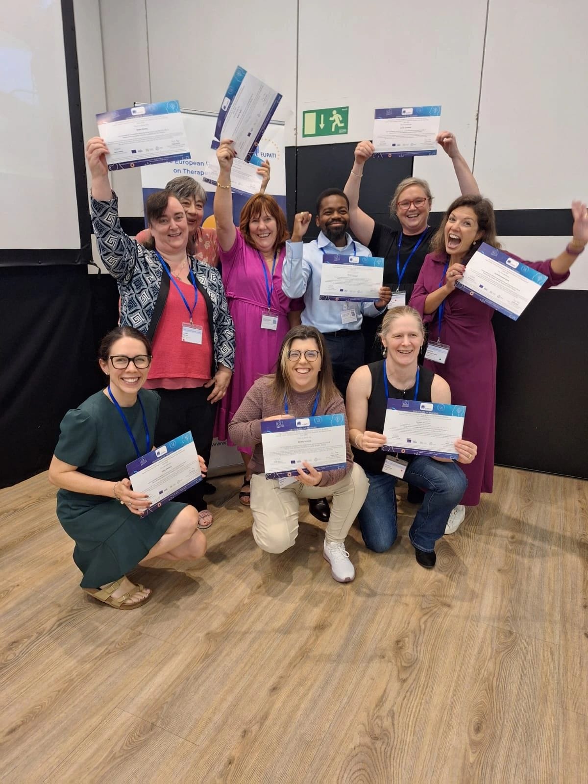 A group of nine people pose indoors, holding certificates and smiling, with some raising their arms in celebration.