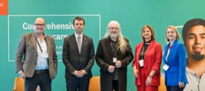 Five people stand in a row on a stage in front of a green backdrop with the text "Comprehensive care." Four chairs and water bottles are visible behind them.