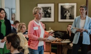 A woman in a coral cardigan speaks to a seated group in a room with green walls and framed photos, while others stand and listen.