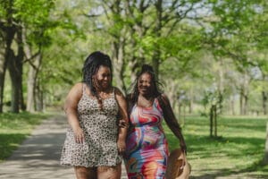 Two women in patterned dresses walk and smile together on a tree-lined park path on a sunny day.