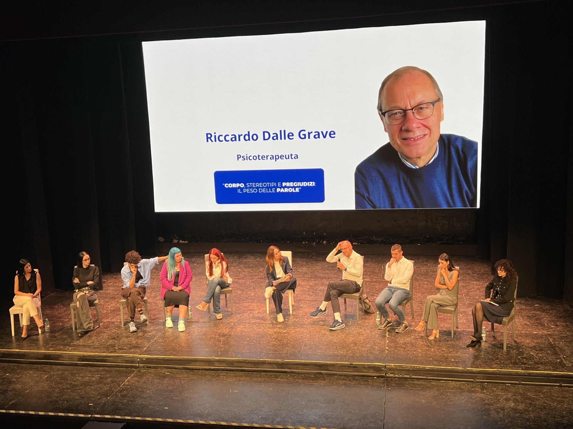 Nine people sit on stage facing the audience, with a large screen behind them displaying a photo and information about Riccardo Dalle Grave, identified as a psychotherapist. Nine people sit on stage facing the audience, with a large screen behind them displaying a photo and information about Riccardo Dalle Grave, identified as a psychotherapist.