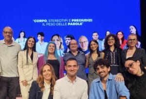 A group of people pose on stage in front of a blue screen displaying Italian text about body, stereotypes, prejudice, and the power of words.