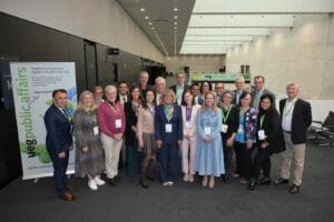 A group of around 25 people pose together in a modern conference hallway, all wearing name badges and green lanyards, with a banner for UEG Public Affairs in the background.