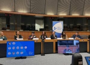 A panel of seven people sits at a long table in a conference room, with screens and a banner displaying "Action on Obesity" in front of them.
