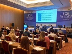 People attend a conference in a hotel meeting room while speakers present slides and discuss on stage during the ECN Masterclass event.