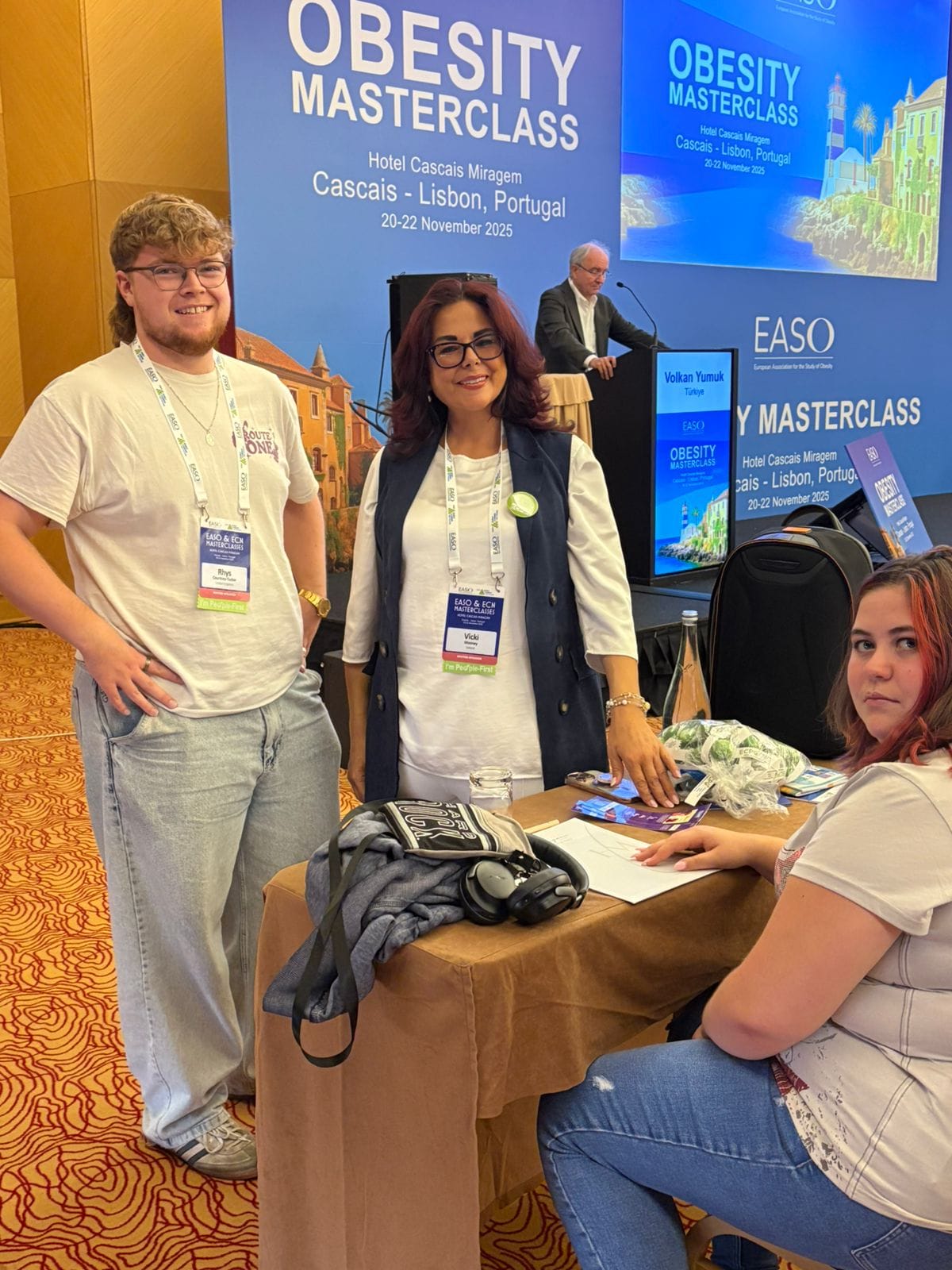 Three people stand and sit at a table with conference materials at the Obesity Masterclass event in Cascais, Lisbon, Portugal. A presentation is displayed on a large screen in the background.